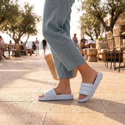 Γυναίκα που περπατά σε ηλιόλουστο, πλακόστρωτο μονοπάτι παραλιακού εστιατορίου, φορώντας ανοιχτό μπλε slides Paredes με φουσκωτό λουρί και γαλάζιο λινό παντελόνι. Κρατά μια ψάθινη τσάντα. Το φόντο δείχνει ελιές και τραπεζοκαθίσματα.
